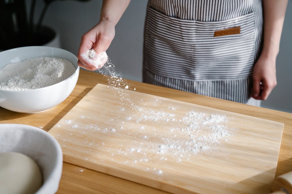 Close-up of a baker sprinkling flour on a wooden board in a kitchen setting.